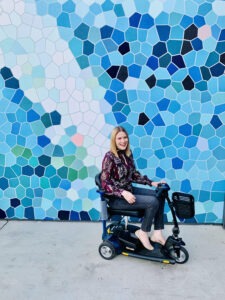 Amy, a disabled, white Australian woman with blonde hair, sits on her mobility scooter in front of a wall with blue and white geometric shapes.