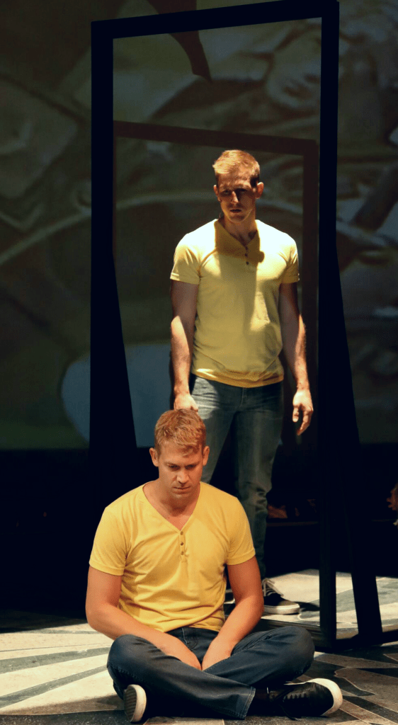 Two men dressed in yellow t-shirts and jeans, are looking down, one standing behind the other