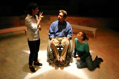 One woman stands, signing, facing a man in a wheelchair and a woman sitting on the floor
